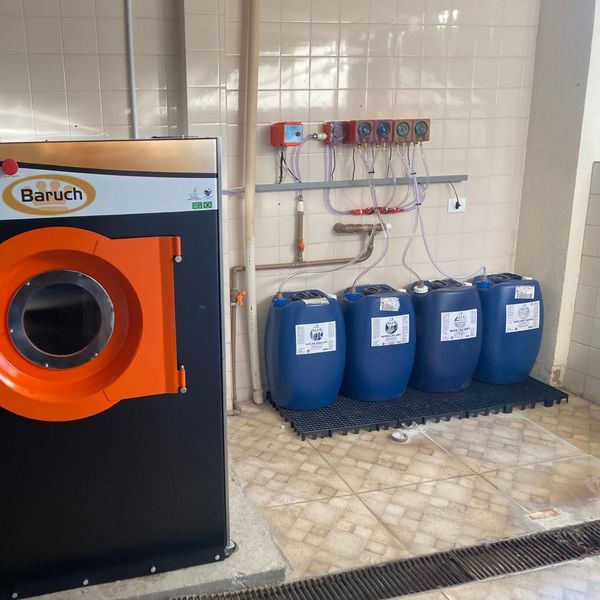 Lavadora industrial Baruch ao lado de quatro bombonas de produto sobre pallet, alimentadas por bombas dosadoras instaladas na parede, em lavanderia atendida pela Protego