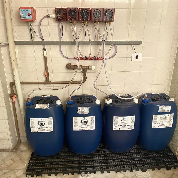 Bombonas Alfa Tex Desinfetante, Alpha Lav ADT, Alfa Tex Oxy e Alfa Soft White ligadas ao painel de bombas dosadoras de uma lavanderia atendida pela Protego